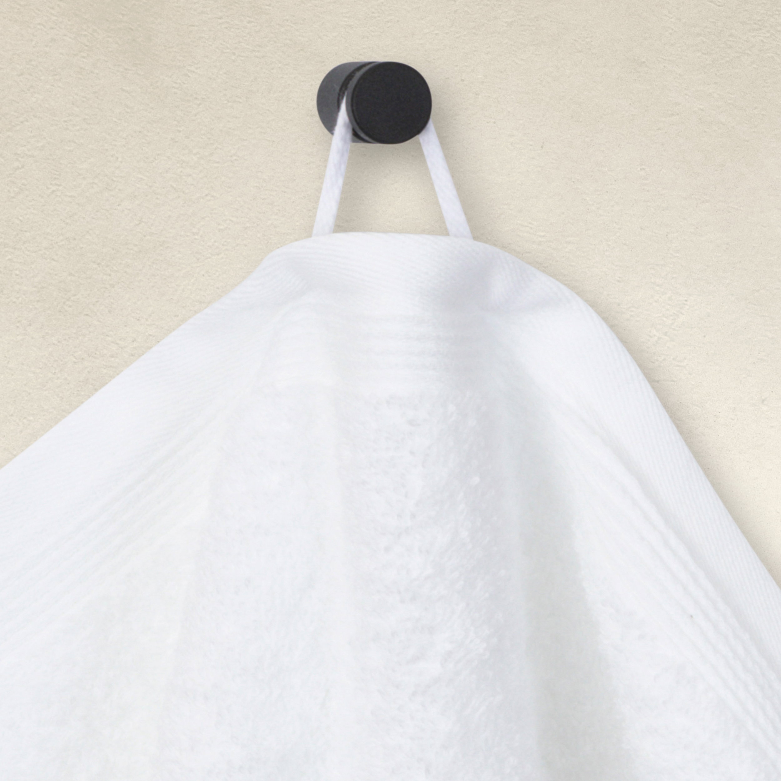 White towel hangs on a round, black wall hook with a hanging loop in front of a light beige, slightly textured wall.