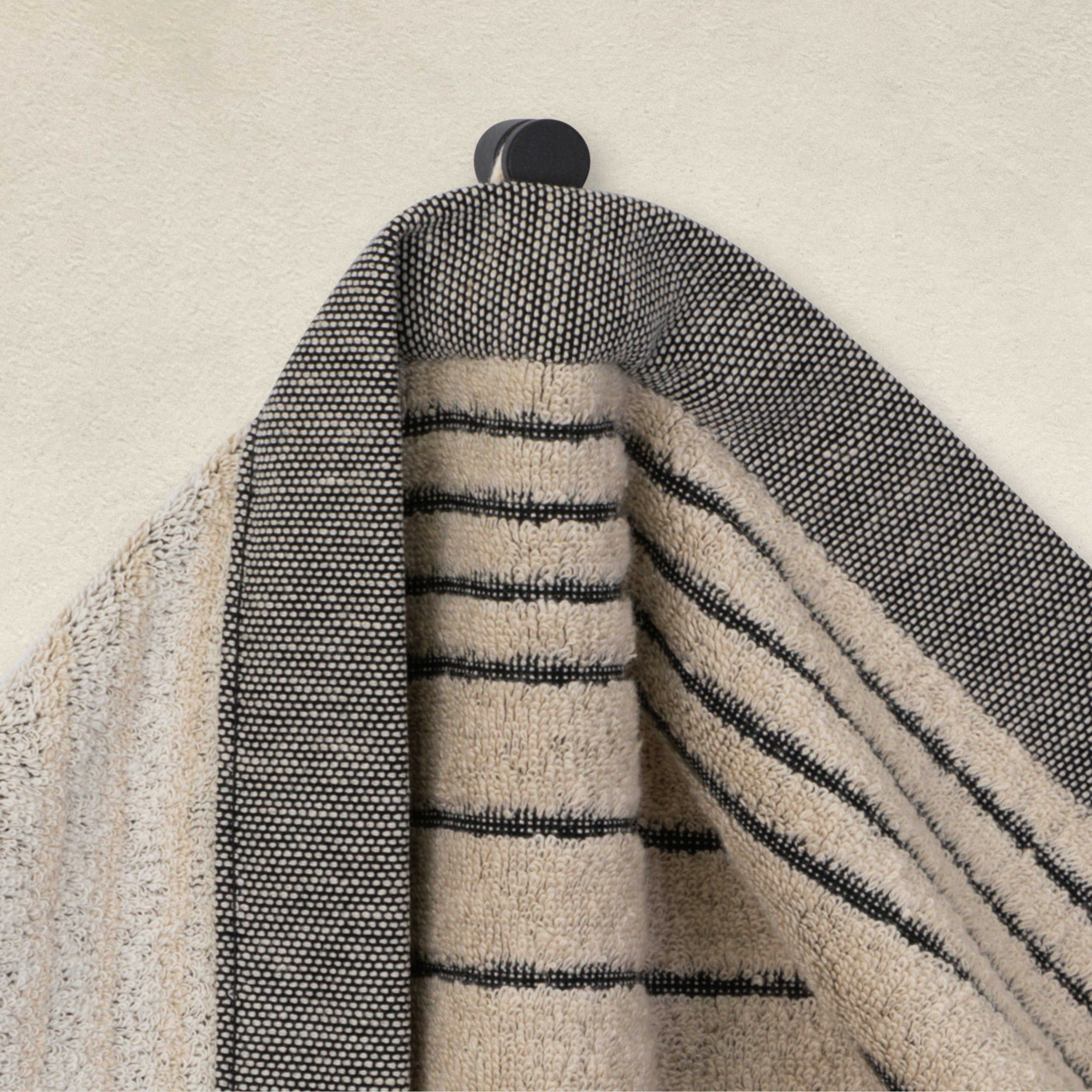 Towel—a beige-and-black-striped bath towel—hangs on a black hook; gray hem and coarse weave visible against a light-colored wall.