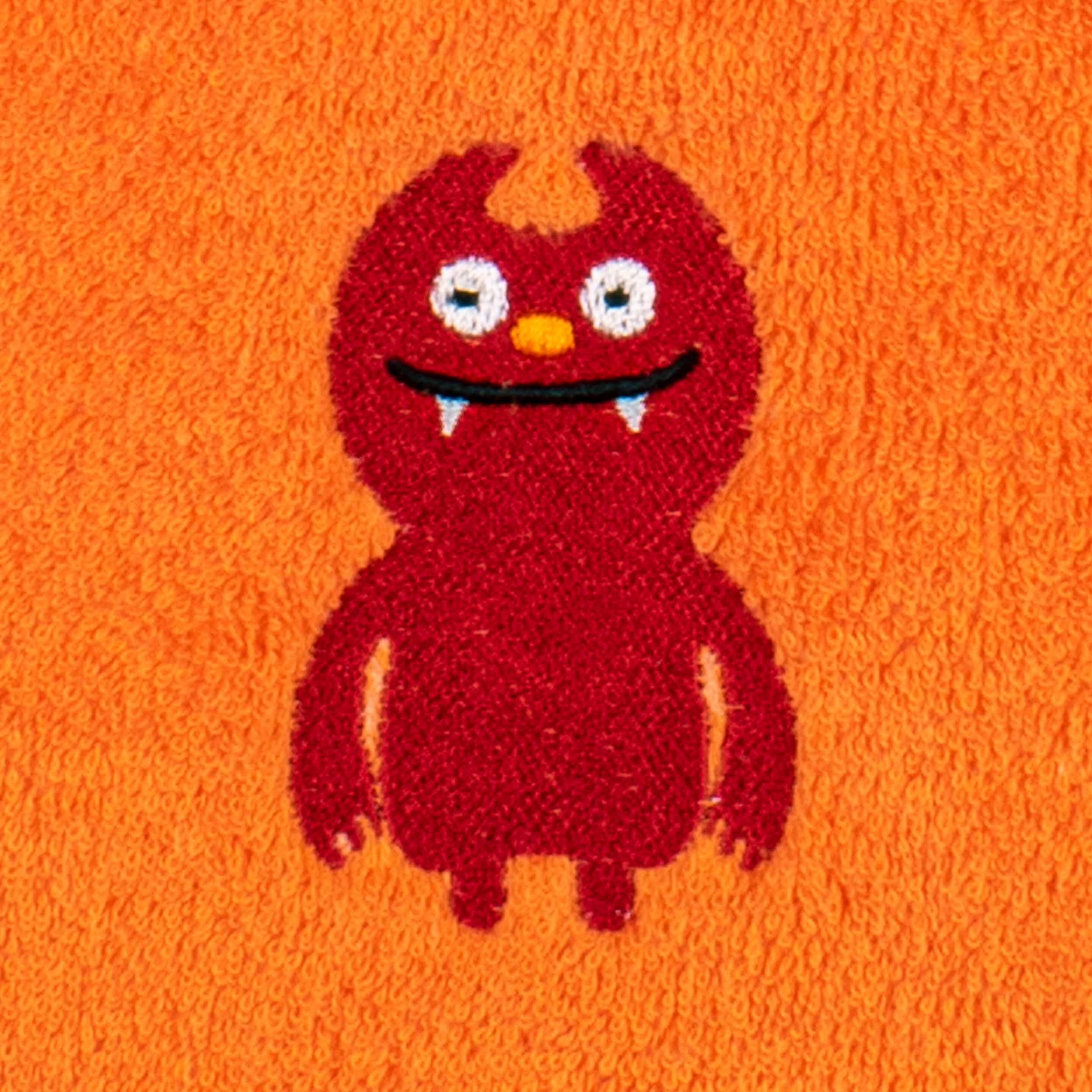 Rotes, cartoonhaftes Monster mit weißen Augen, gelber Nase und zwei Fangzähnen ist als fröhliche Stickerei auf einem orangefarbenen Frottee-Badetuch platziert.