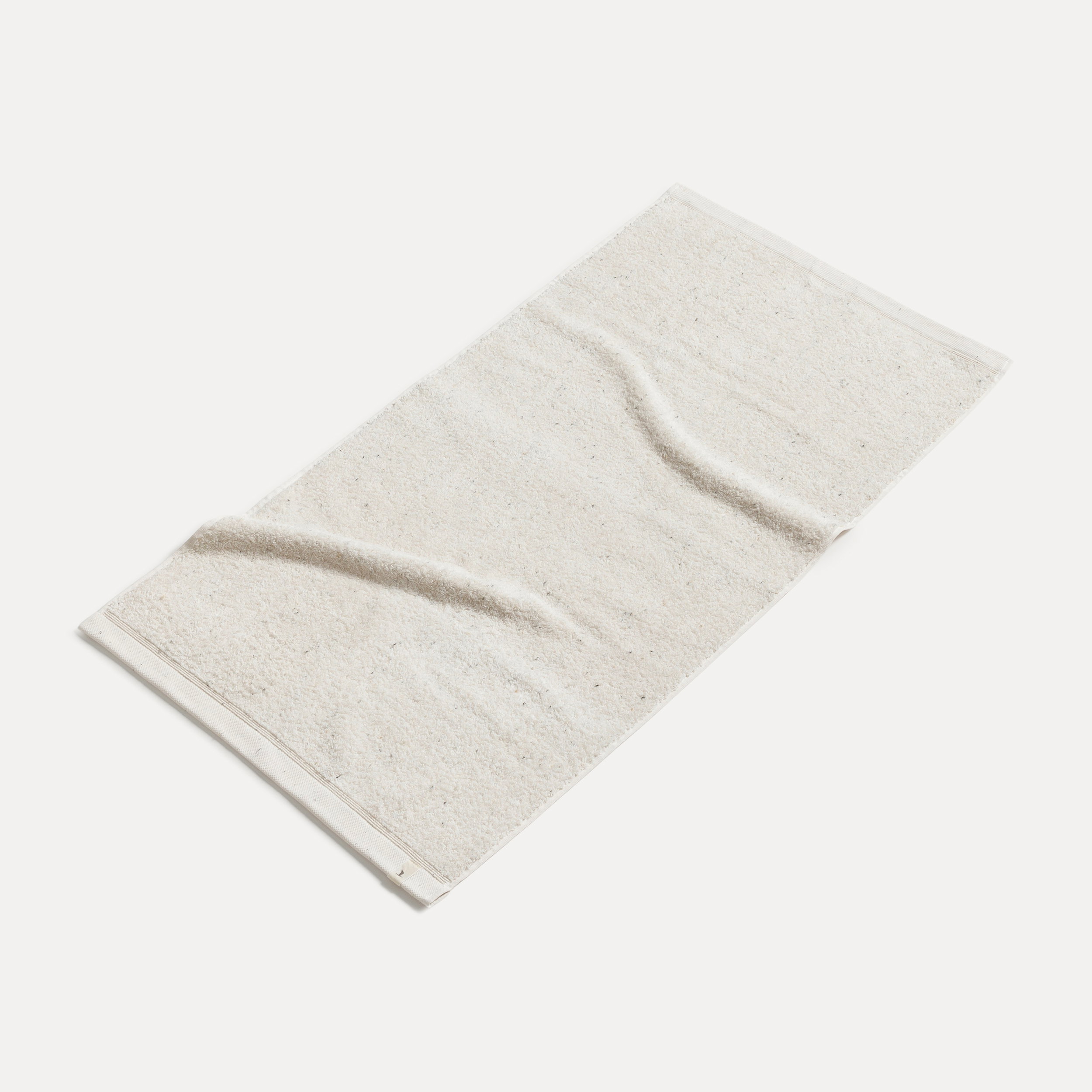 A light beige towel lies slightly crumpled and flat on a light background, showing the fabric texture and hem, and presenting a simple bathroom accessory.