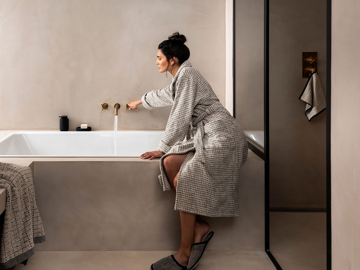Person im Bademantel dreht den Wasserhahn einer Badewanne auf; beiges, minimalistisches Badezimmer mit Handtüchern und Glasduschwand.