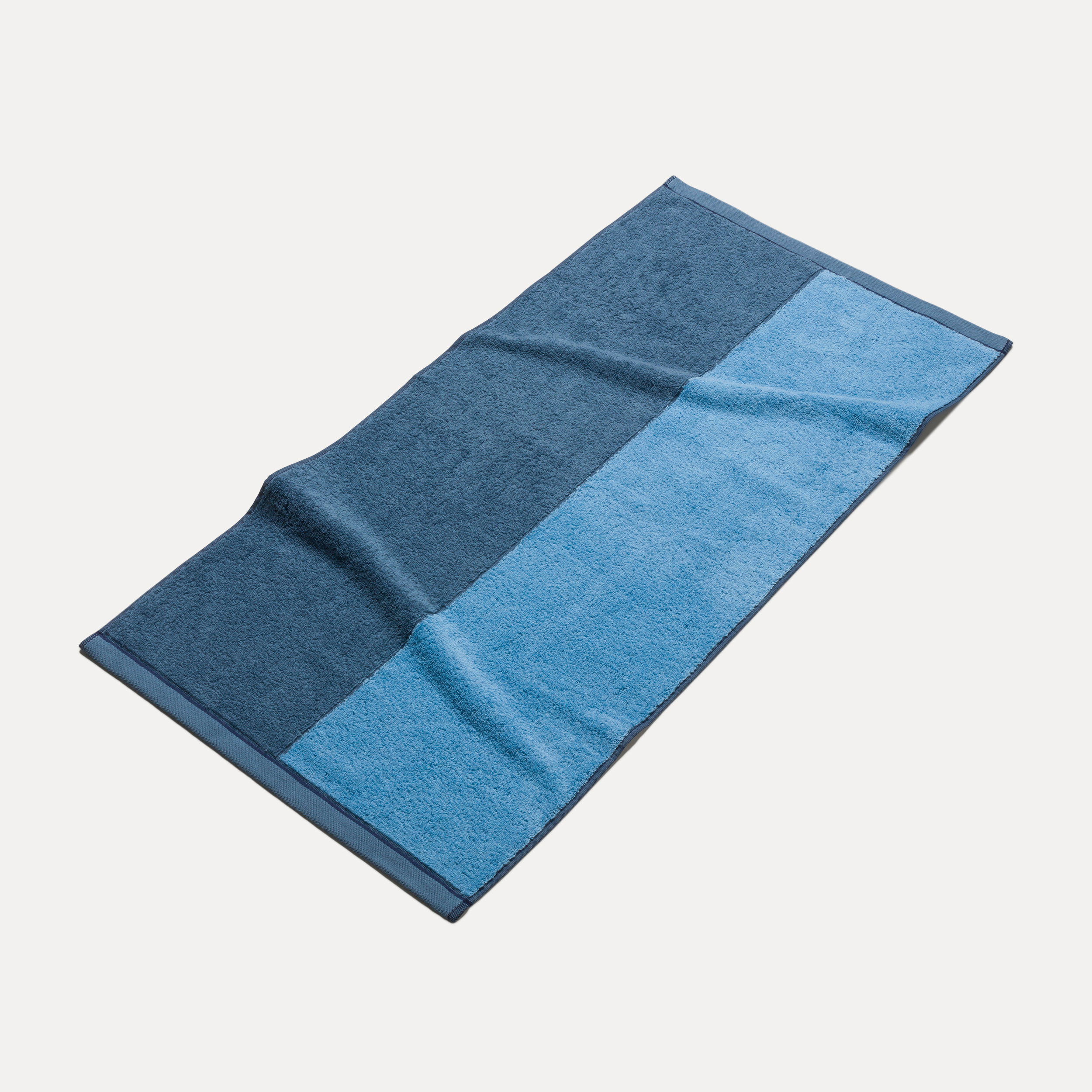 Two-tone bath towel in dark and light blue lies flat, slightly rippled; isolated against a light, neutral background.