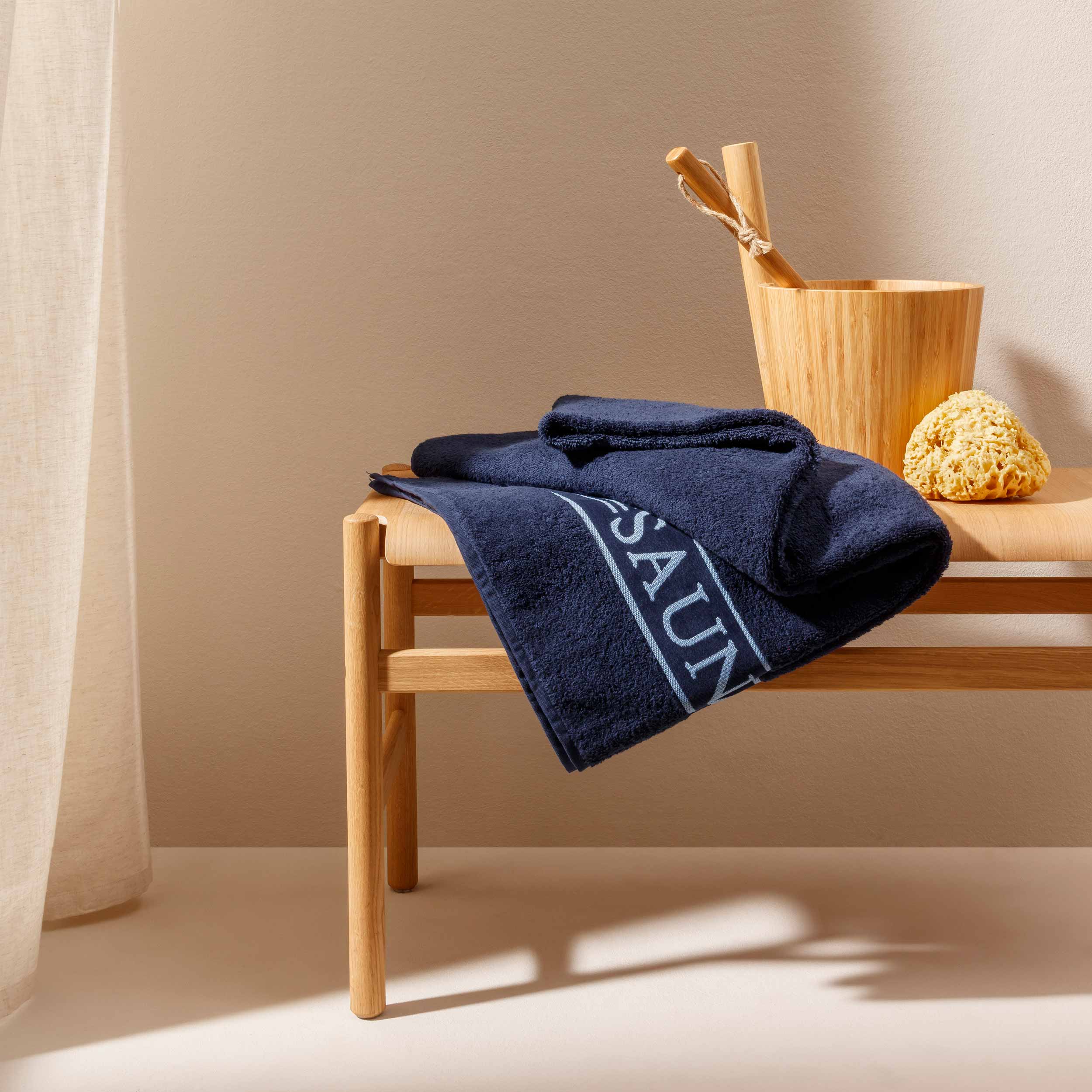 Dunkelblaue Handtücher liegen gefaltet auf einer hellen Holzbank; daneben ein Bambus-Saunakübel mit Kelle und ein Natur-Schwamm, vor beiger Wand und leichtem Vorhang., Handtuch, Navy, Badehandtuch, Holzbänken, Badezimmer-Dekoration,  Handtuch, Badetuch, m