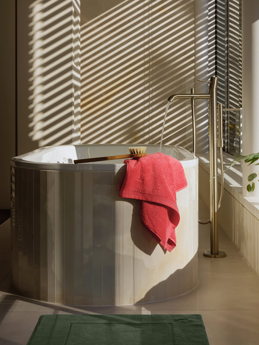A pink towel hangs over a round bathtub; a brush lies on top of it. A bright room with a freestanding faucet, a green bath mat, and the shadows of blinds in the sunlight.