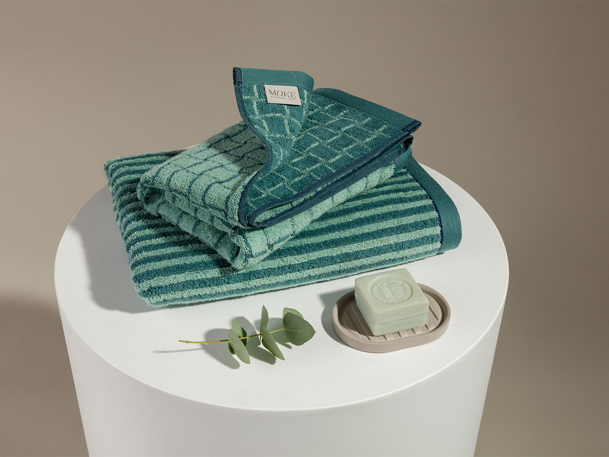 Folded green-turquoise towels stacked on a round white pedestal; next to it, a soap dish with a bar of soap and a eucalyptus branch against a neutral beige background.