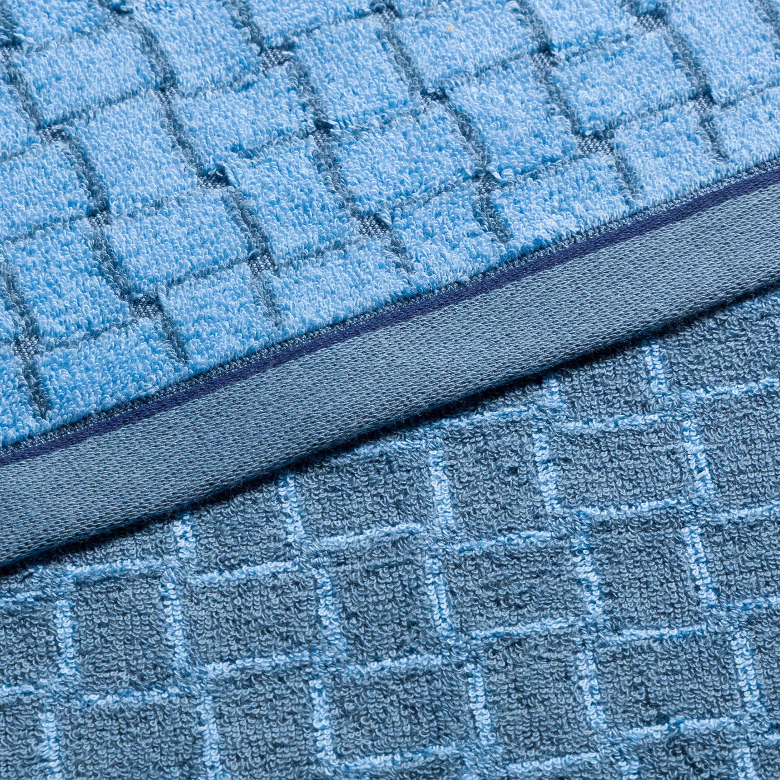 Two light and dark blue terry cloth towels with a checkered pattern, stacked on top of each other and folded over at the edge; close-up, emphasizing texture and material, neutral background.