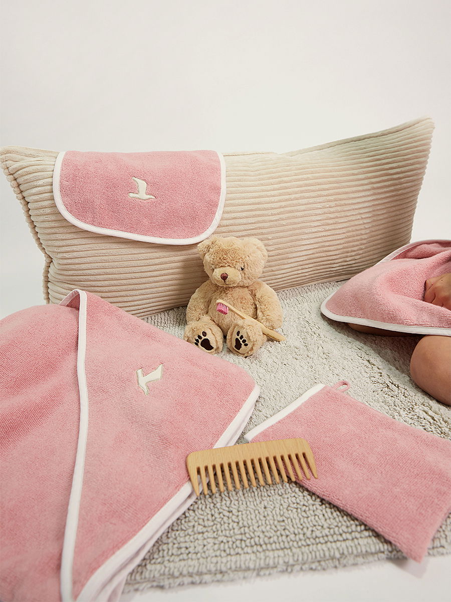 Pink terry cloth items (hooded towel, bath towel, washcloth) are arranged neatly; a small teddy bear sits in the center, and a wooden comb lies in front on a gray bath mat in front of a striped pillow.