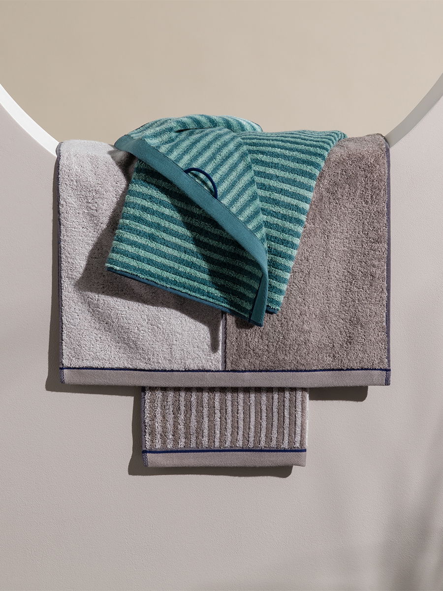 Several Möve towels (beige, gray, and turquoise-striped) are draped over each other and hung on a semicircular metal ring; a small guest towel hangs below, neutral wall background.