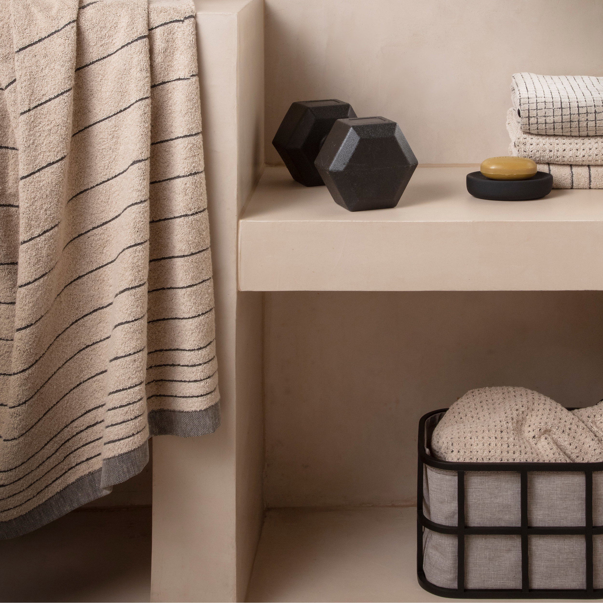 A beige towel with fine dark stripes hangs over a light-colored stone shelf; next to it, a soap dish with yellow soap, two black hexagonal objects, folded towels, and a metal basket.