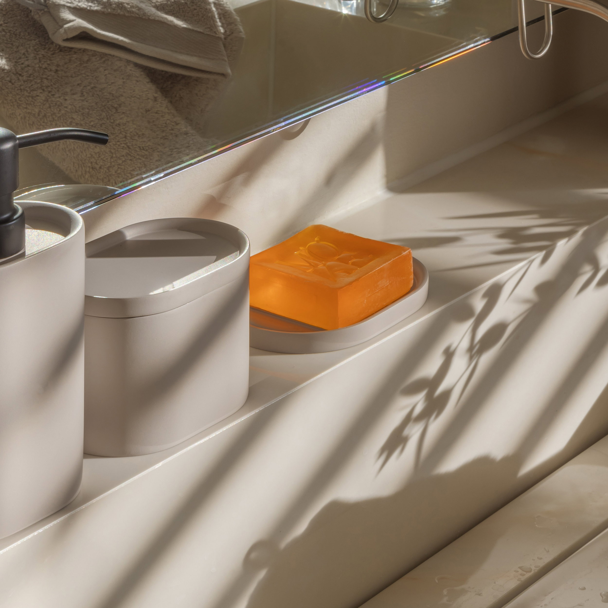 Orangefarbenes Seifenstück liegt auf einer Seifenschale, umgeben von Seifenspender und Behälter; Sonnenlicht und Pflanzenschatten fallen auf den Waschbeckenrand vor einem Spiegel., Seife, Orangene Seife, Waschraum, Soap bar, Küchen/accessoire