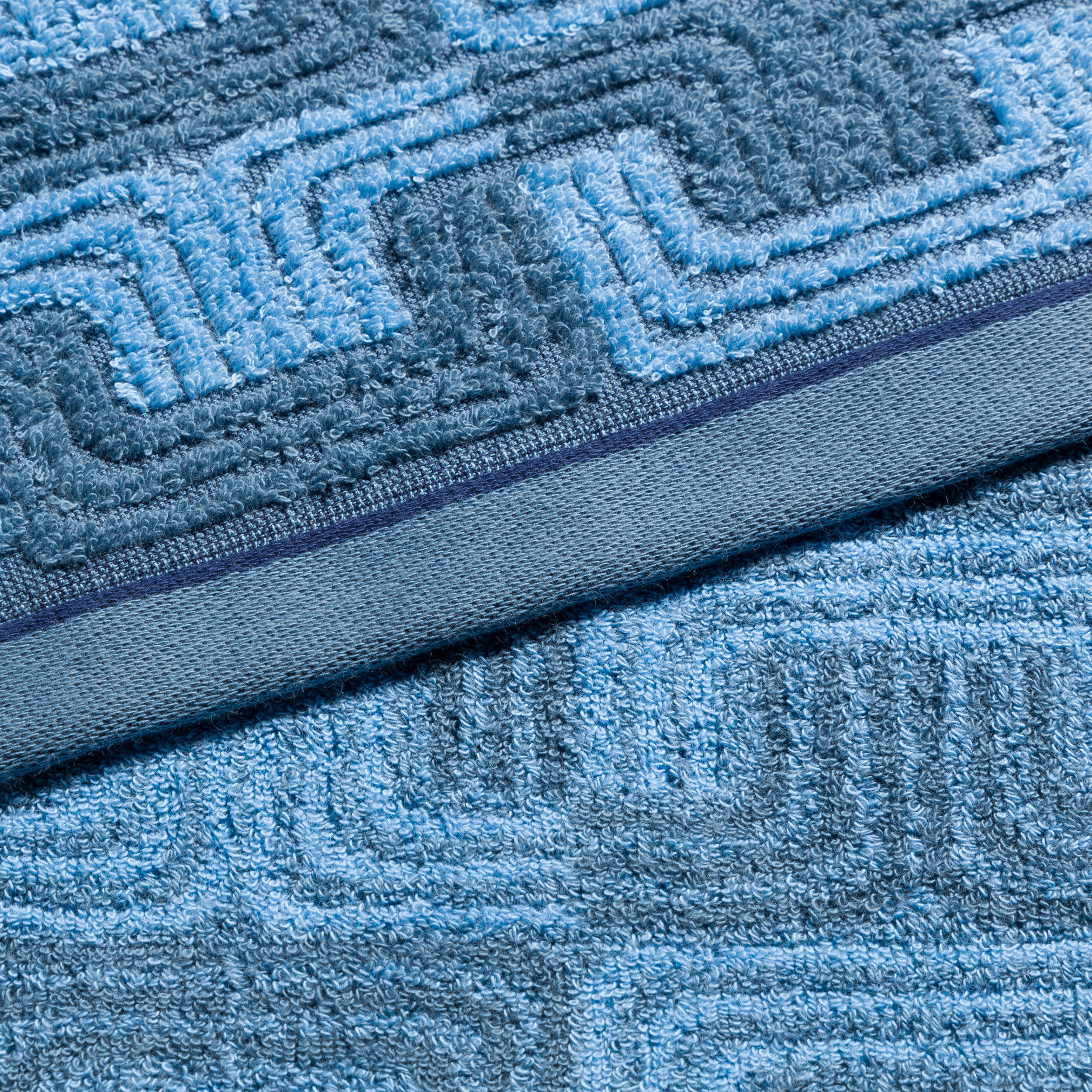Ein blaues Handtuch liegt gefaltet, zeigt ein reliefiertes, geometrisches Linienmuster; Nahaufnahme betont weiche Frotté-Textur und schmale dekorative Bordüre als Badezimmeraccessoire.