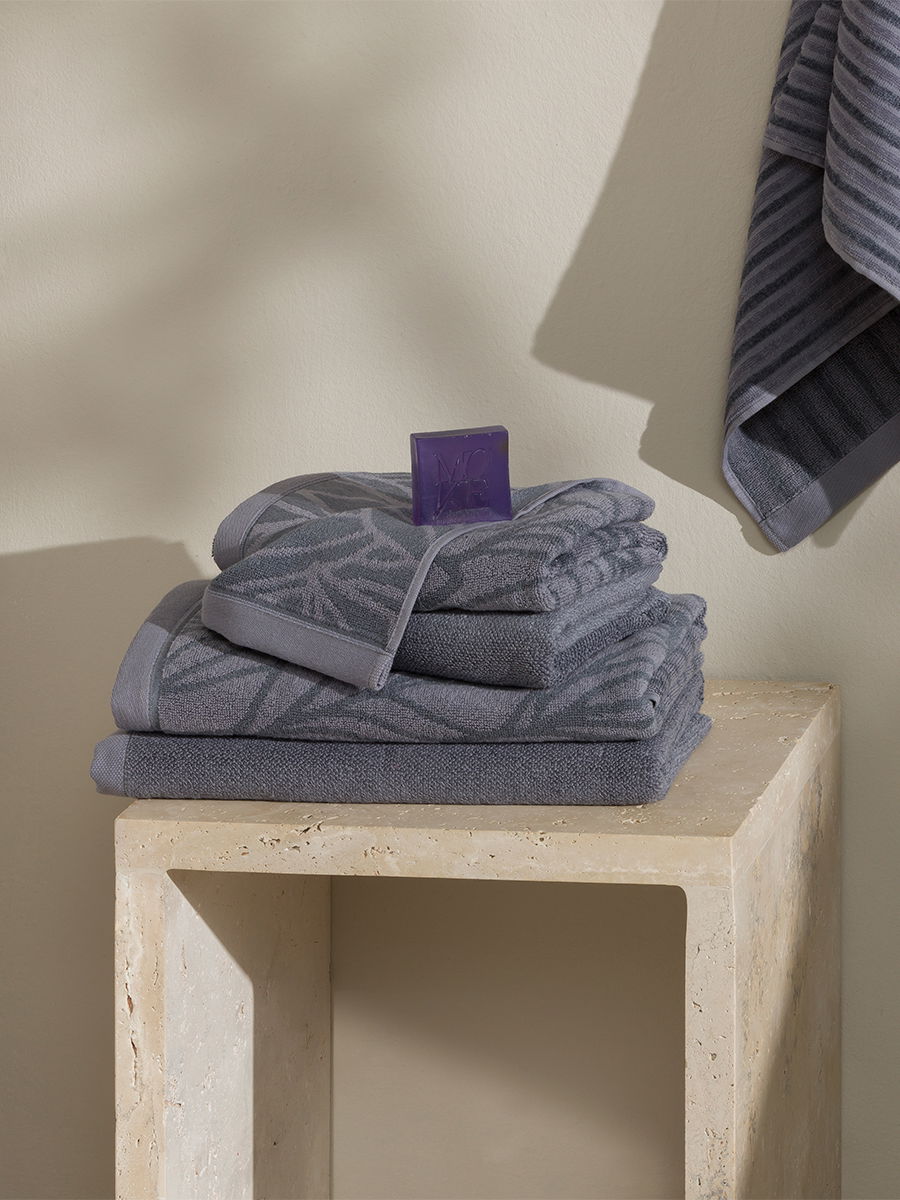 A stack of gray-blue, patterned towels with purple loops, neatly folded, rests on a light-colored, rectangular stone pedestal; in the background, another towel hangs on the wall.