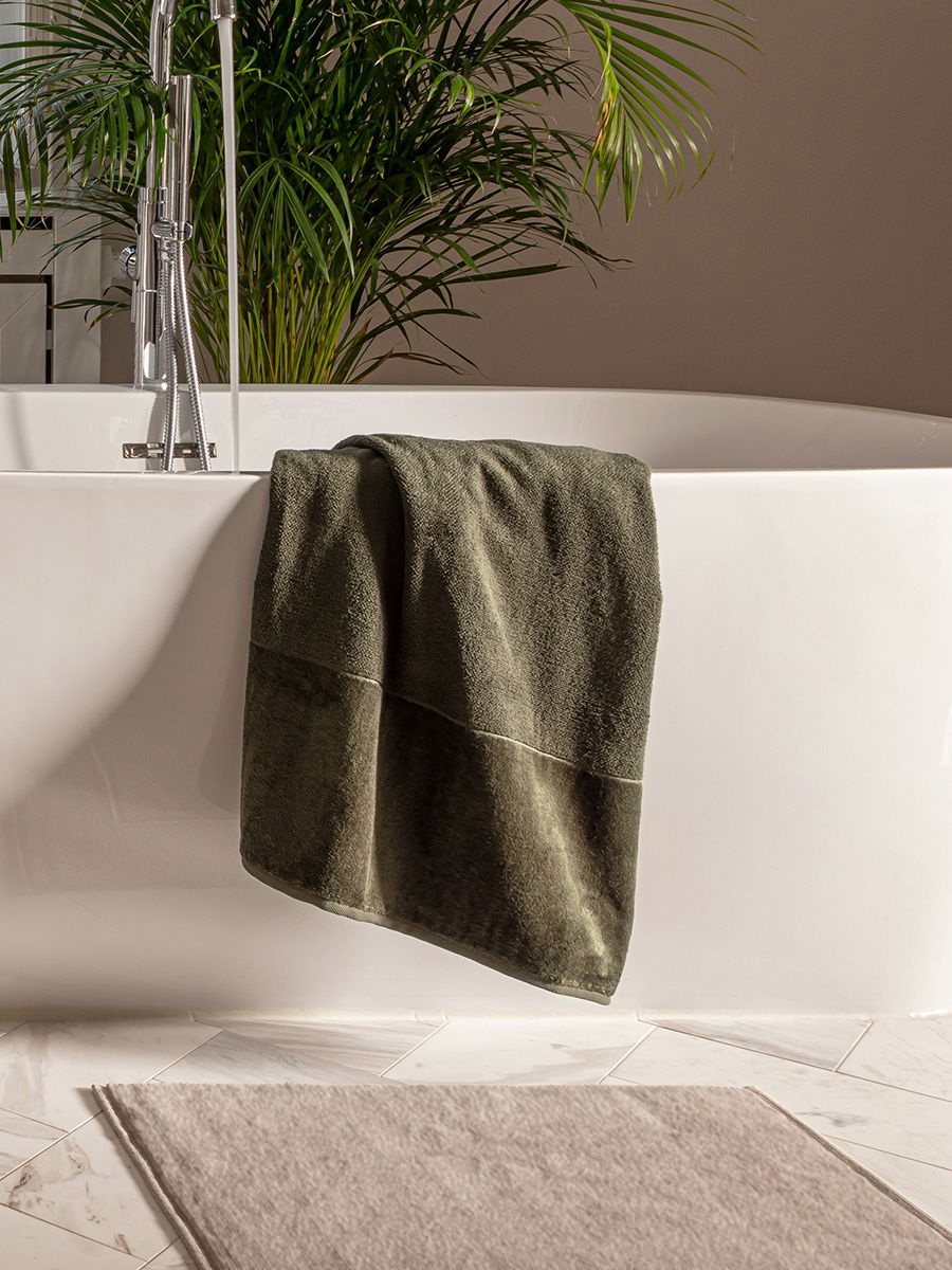 Dunkelgrünes Handtuch hängt über den Rand einer weißen freistehenden Badewanne; ruhiges Badezimmer mit beiger Wand, großer Zimmerpflanze im Hintergrund und heller Badematte auf Fliesen.