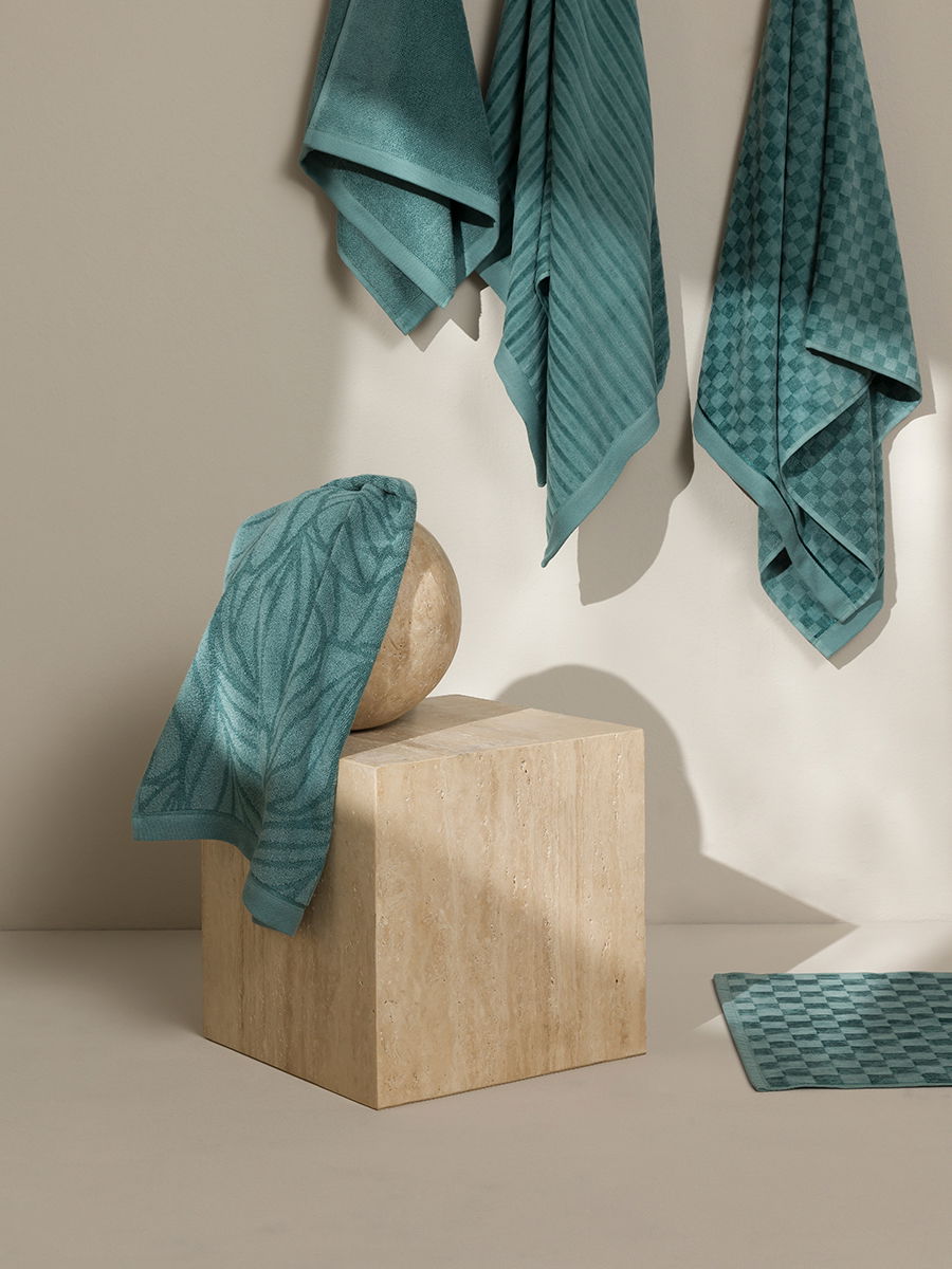 Several turquoise-blue towels hang on the wall; one covers a sphere on a wooden block; a bath mat lies on the floor — a minimalist, studio-like bathroom setting.