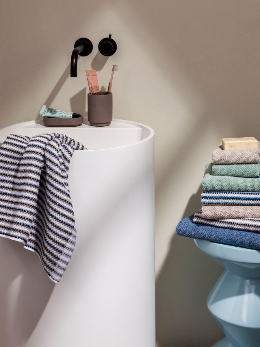 Handtücher: ein gestreiftes hängt über einem weißen Säulenwaschbecken, weitere gefaltet auf einem blauen Hocker; Zahnbürsten und Seife neben schwarzem Wandhahn, schlichtes Badezimmer.