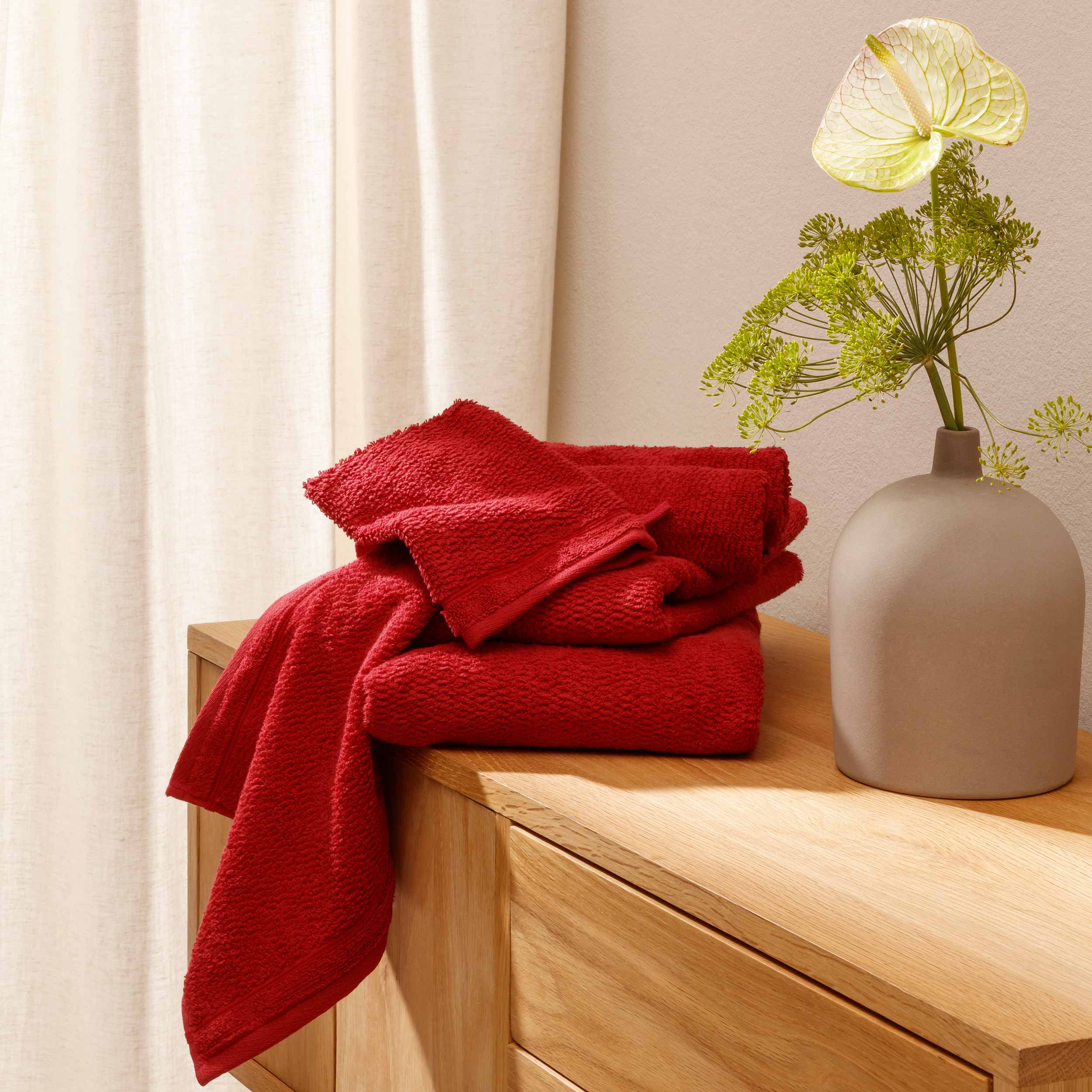 Rote, gefaltete Handtücher liegen gestapelt und teils herabhängend auf einer hellen Holzkommode; rechts steht eine matte Vase mit grünen Blüten, im Hintergrund ein cremefarbener Vorhang., Handtücher, Rotes Handtuch, Badetuch, Baumwolltuch, Wohnkultur, Woh