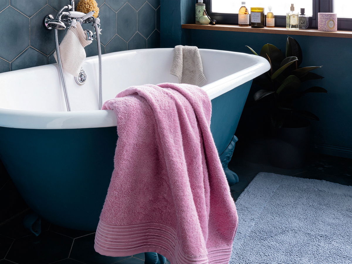 Ein rosa Handtuch liegt drapiert über dem Rand einer freistehenden Badewanne; im Hintergrund weiteres helles Handtuch, dunkelblaue Wände/Fliesen, Fensterbrett mit Pflegeprodukten und Pflanze.