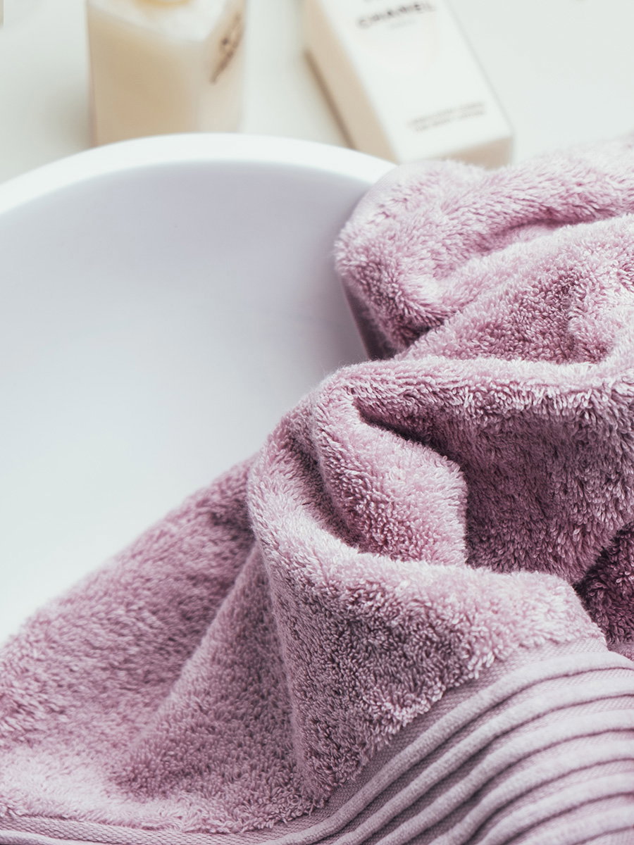 A pink, fluffy towel lies loosely in a white bowl; out-of-focus toiletries on a light surface in the background, simple bathroom arrangement.