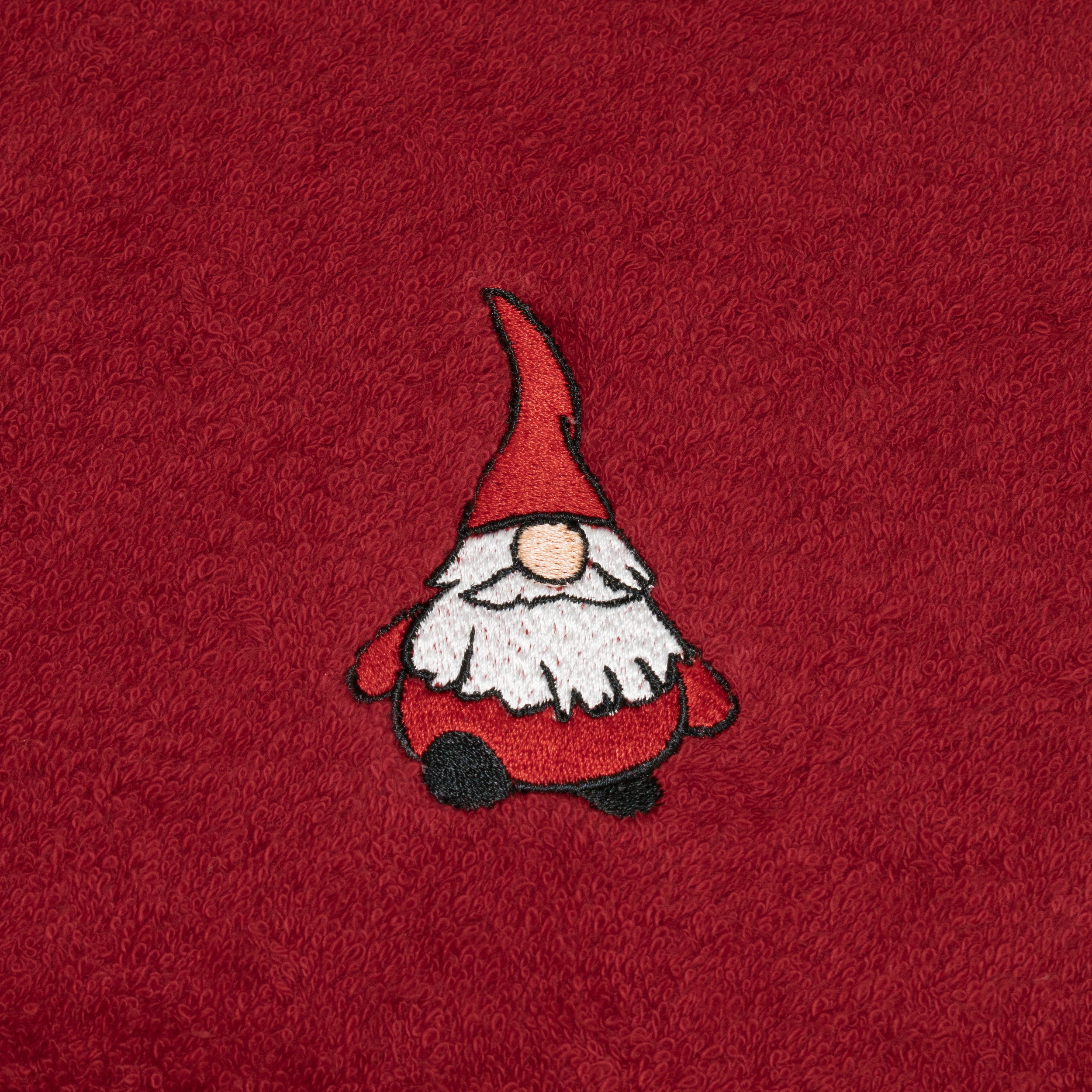 An embroidered elf sits decoratively on a red terry cloth towel; white beard, red hat and body, black shoes, visible embroidery pattern as a decorative bathroom accessory.