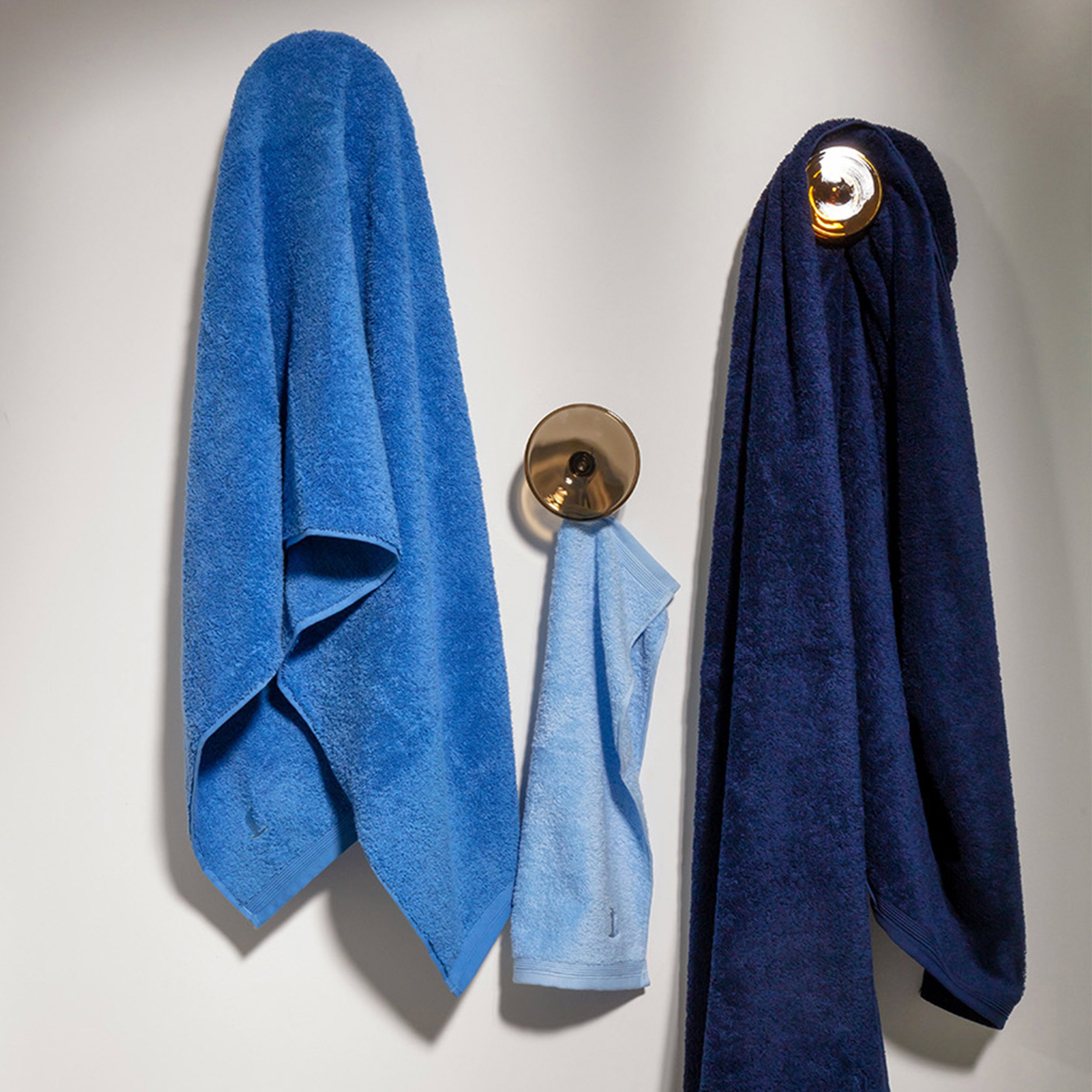 Three towels (dark blue, blue, light blue) hang loosely on gold-colored hooks on a light-colored wall; simple bathroom arrangement.