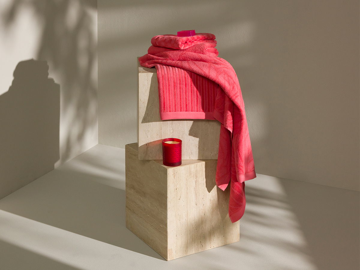 Pink towel and bathrobe, loosely draped over two light-colored stone pedestals; small red vessel next to them; minimalist, sun-drenched room with striking shadows.