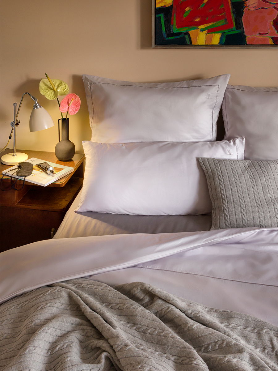 Bett mit hellgrauen Kissen und seidig schimmernder Bettwäsche — Kissen geschichtet; gestrickte Decke lässig darüber; Nachttisch mit Lampe, Vase und Buch, farbiges Bild an der Wand.