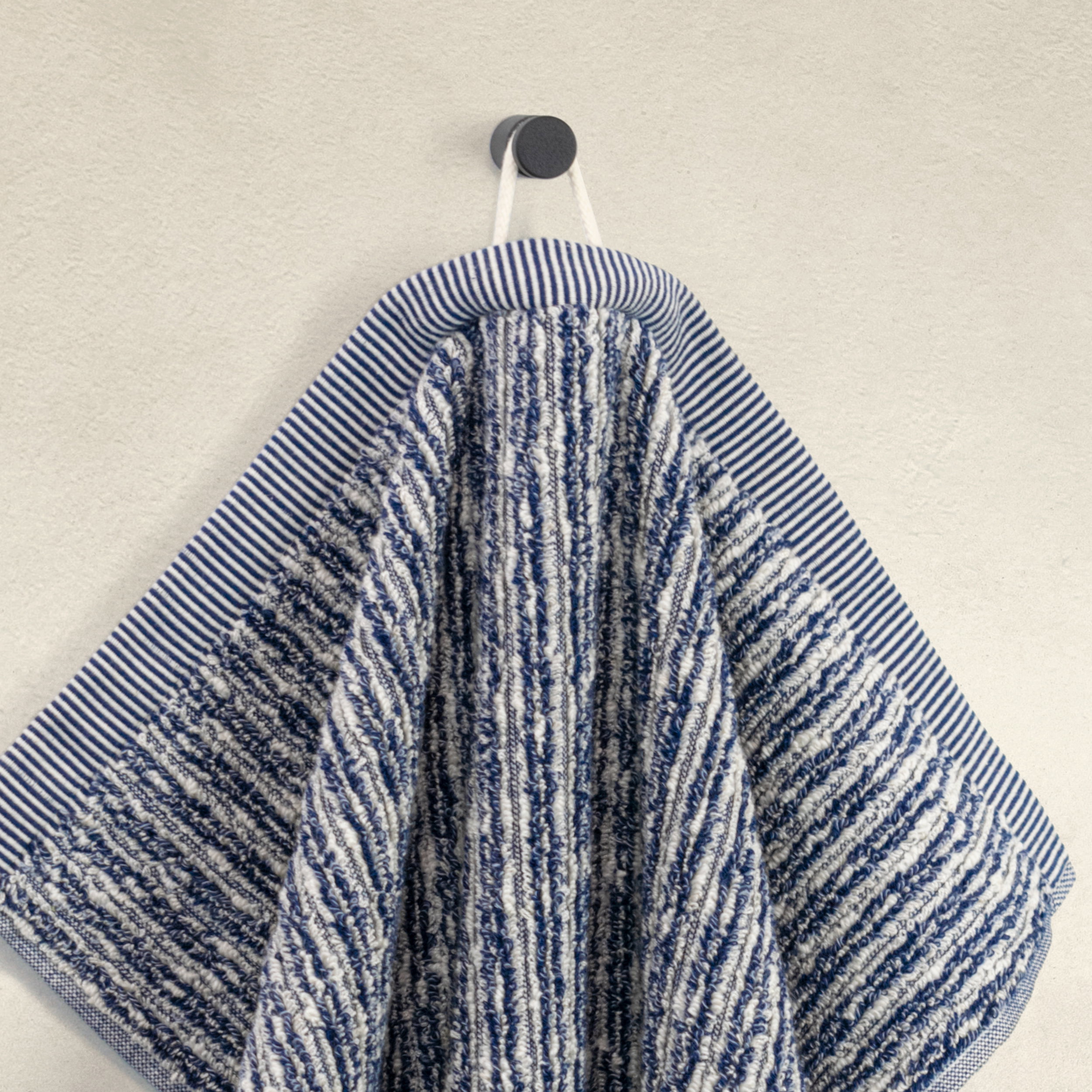 Ein blau-weiß gestreiftes, strukturiertes Handtuch hängt leicht gefaltet an einem runden Wandhaken vor einer glatten, hellen Wand.
