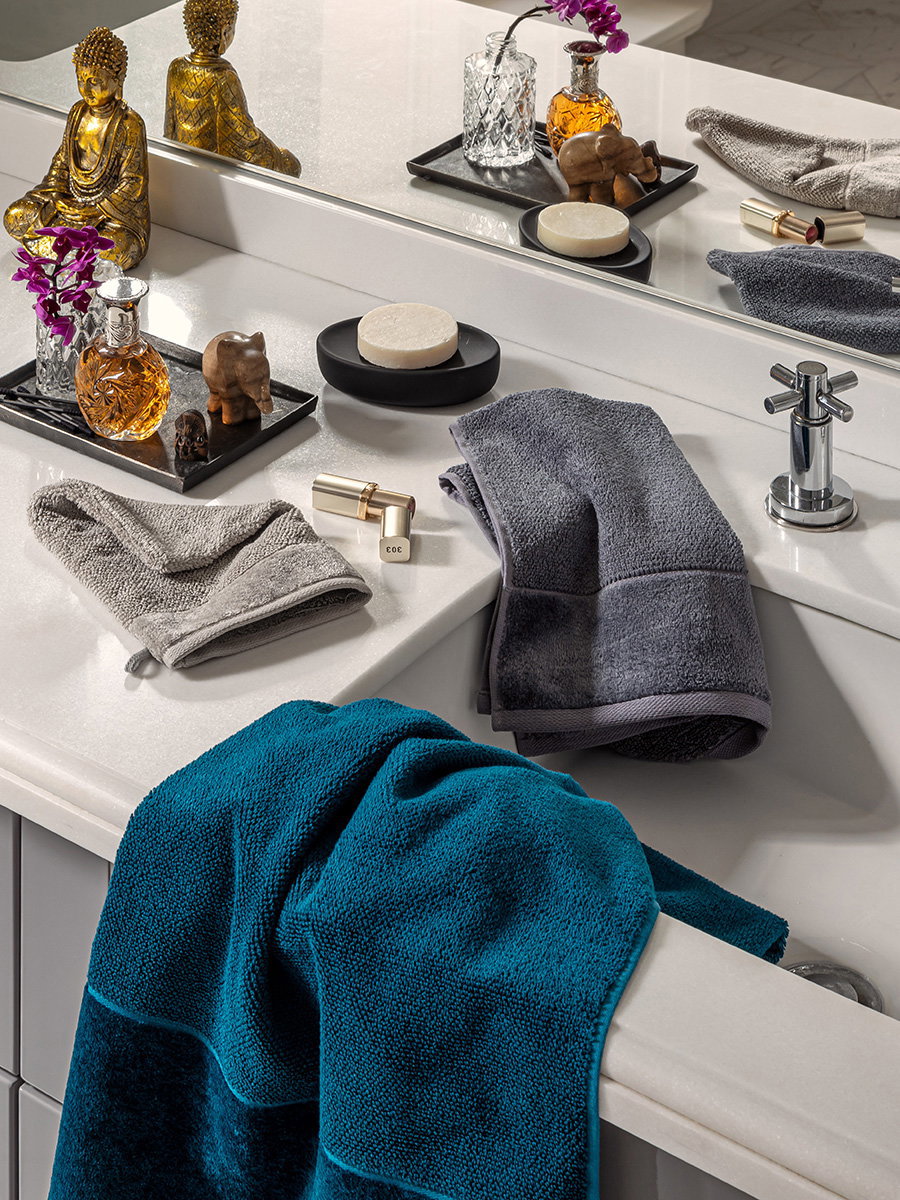 Towels in turquoise and gray tones draped over the edge of a sink; soap dish, perfume bottle, and golden decorative figurine in front of a mirror in a bright, modern bathroom.