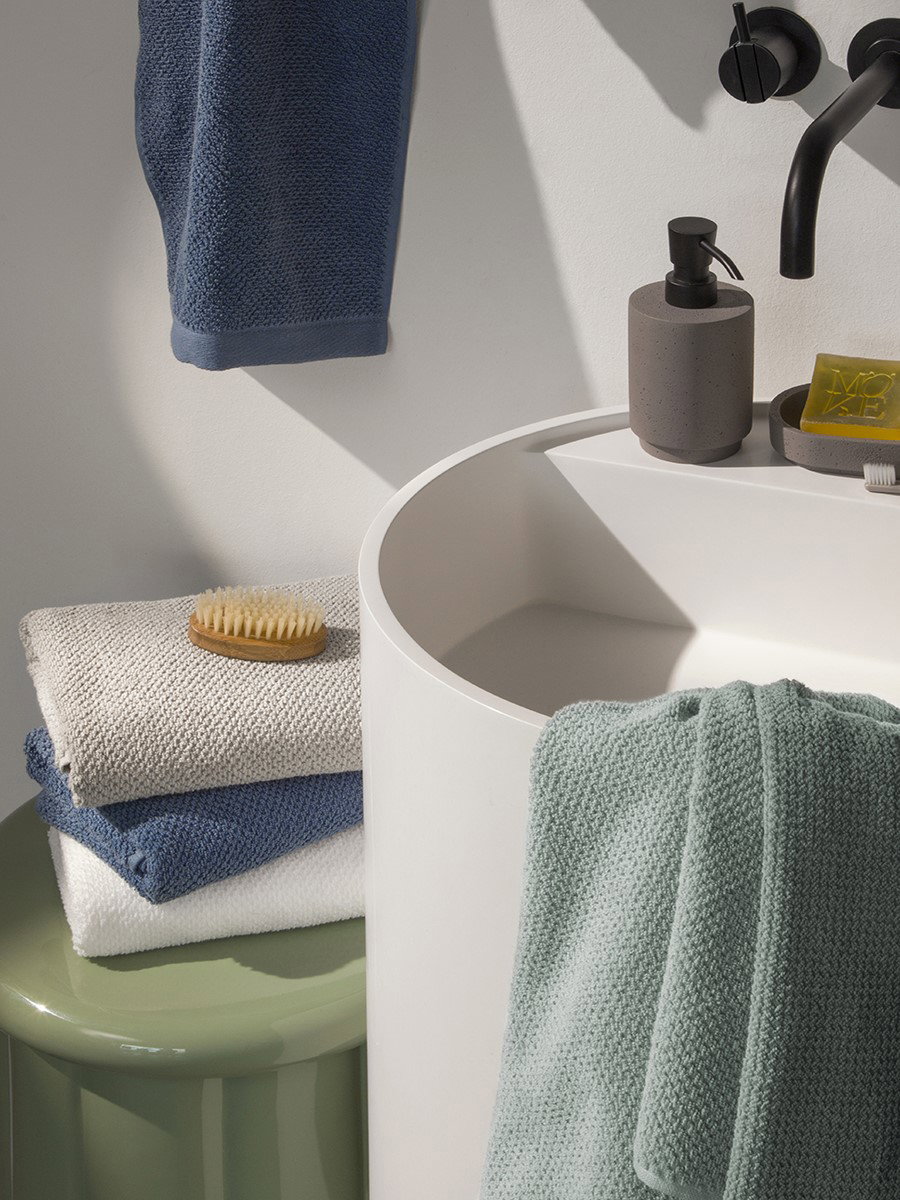 A mint-green, textured towel hangs over the edge of a round white sink; a soap dispenser and soap sit on the sink rim, next to folded towels with a brush on a green stool, against a light-colored wall.