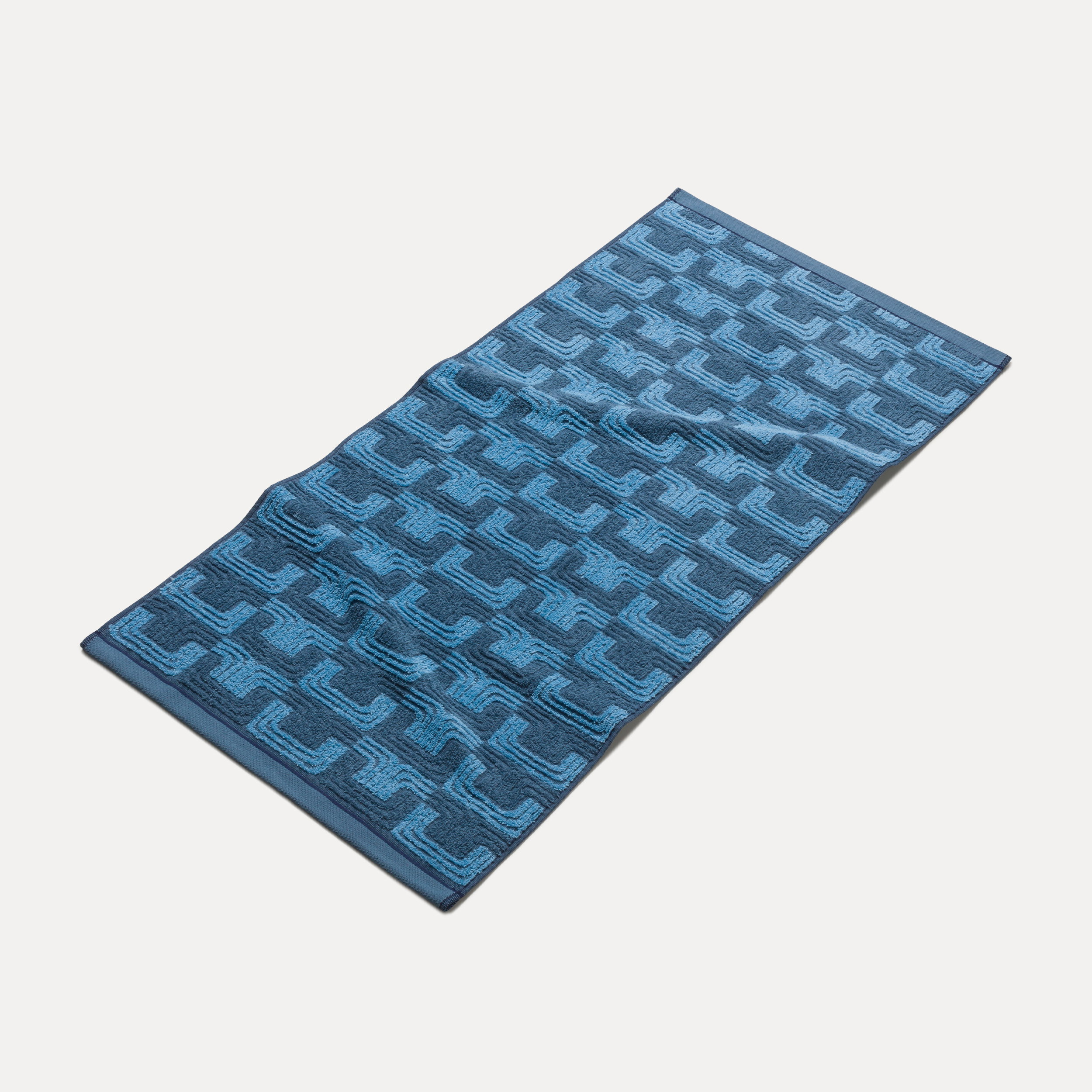 Handtuch liegt flach ausgebreitet; blaues Badetuch mit geometrischem, gestreiftem Muster in verschiedenen Blautönen, leicht zerknittert, auf neutralem hellgrauem Hintergrund präsentiert.