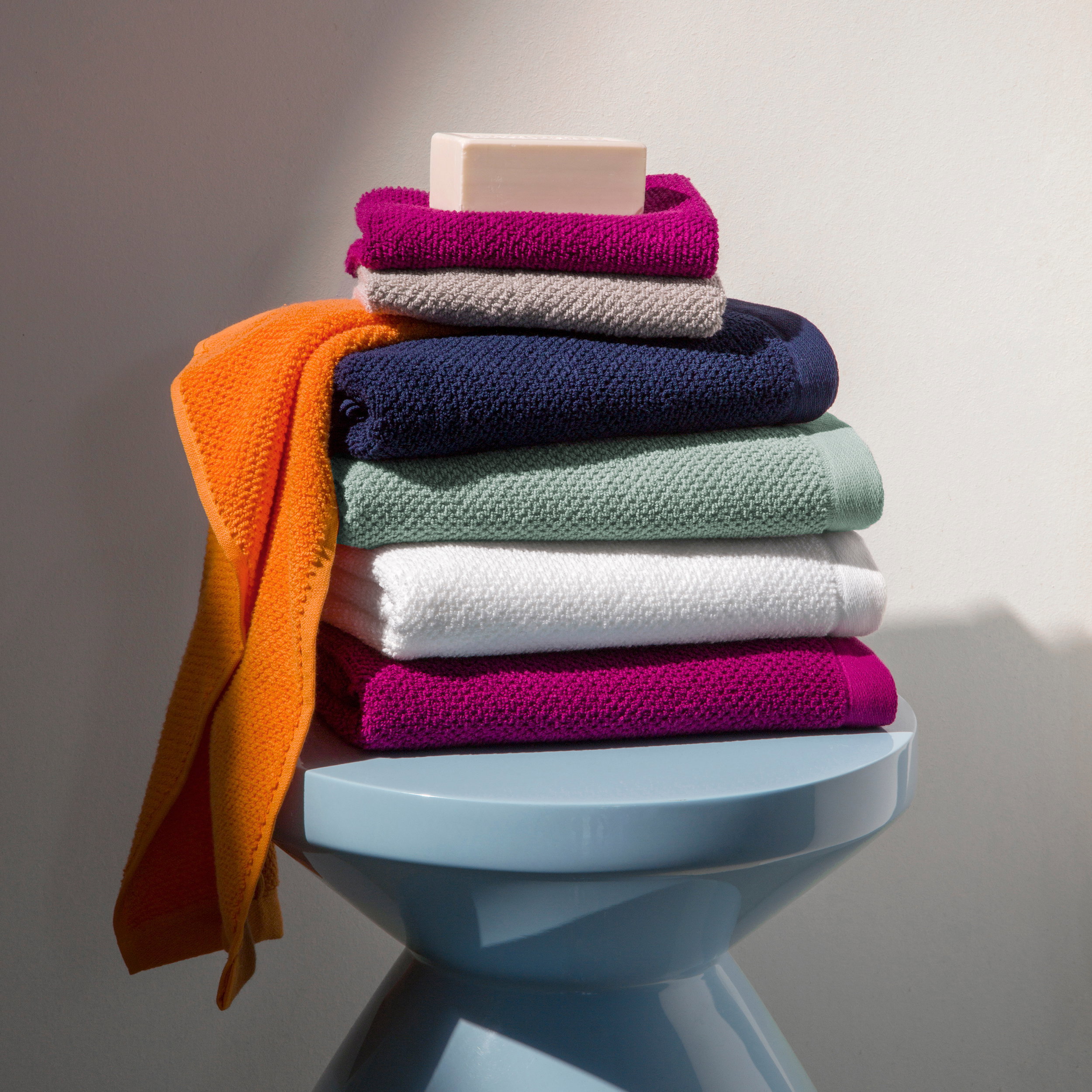 A stack of colorful towels is piled on a light blue stool, with a bar of soap on top, against a neutral, light background.